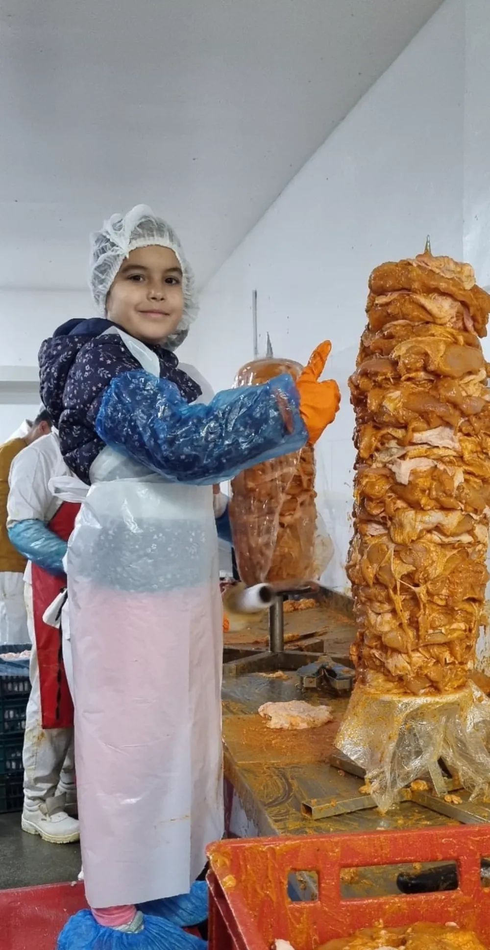 O fetiță cu bonetă și mănuși de protecție ridică degetul mare lângă un rulou de shaorma, în hala de producție.